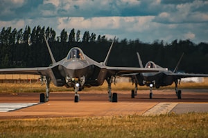 Two sleek, modern fighter jets are taxiing on an airstrip surrounded by grassy fields. The lead jet is prominently visible in the foreground, showcasing its angular and stealthy design against a backdrop of trees and a partly cloudy sky. The second jet follows behind at a distance.