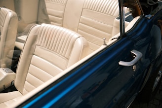 A classic car interior with beige leather seats, visible through the open door of a blue vintage vehicle. The seats feature vertical stitching patterns and a sleek design. The door handle and locking mechanism are visible on the car door.