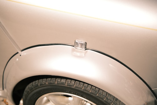 A close-up view of a car's wheel and fender area. The tire is visible with a tread pattern, mounted on a shiny rim. Above the wheel, there is a clear car indicator light attached to the smooth, metallic surface of the car body.