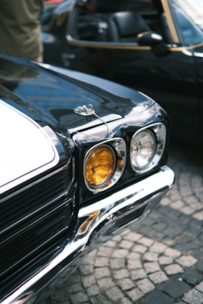 A close-up view of a vintage car's front area, highlighting its headlight assembly and chrome bumper. The headlight has both clear and amber lights, adding to the classic look. The car's surface is polished and reflects the surrounding environment. Another similar car is partially visible in the background.
