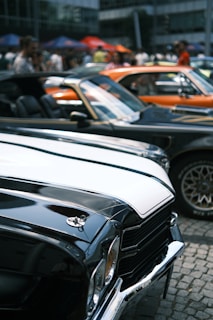 A group of classic cars is lined up on a cobblestone street. The cars have polished exteriors, and their vibrant colors shine under the daylight. People are gathered in the background, some standing under colorful tents. The scene suggests a lively car show or an outdoor event.