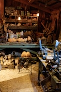 A cluttered workshop filled with woodworking tools and equipment. Wooden clogs are prominently displayed, some resting on a stool in the center. Shelving is packed with various items, including cans, jars, and wooden molds. The floor is covered in sawdust, adding to the rustic atmosphere. Overhead lighting provides a warm glow to the scene.