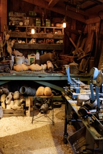 A cluttered workshop filled with woodworking tools and equipment. Wooden clogs are prominently displayed, some resting on a stool in the center. Shelving is packed with various items, including cans, jars, and wooden molds. The floor is covered in sawdust, adding to the rustic atmosphere. Overhead lighting provides a warm glow to the scene.
