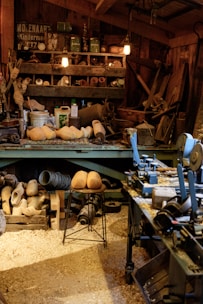 A cluttered workshop filled with woodworking tools and equipment. Wooden clogs are prominently displayed, some resting on a stool in the center. Shelving is packed with various items, including cans, jars, and wooden molds. The floor is covered in sawdust, adding to the rustic atmosphere. Overhead lighting provides a warm glow to the scene.