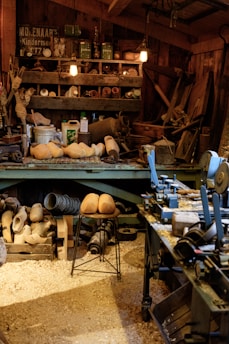 A cluttered workshop filled with woodworking tools and equipment. Wooden clogs are prominently displayed, some resting on a stool in the center. Shelving is packed with various items, including cans, jars, and wooden molds. The floor is covered in sawdust, adding to the rustic atmosphere. Overhead lighting provides a warm glow to the scene.