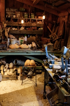 A cluttered workshop filled with woodworking tools and equipment. Wooden clogs are prominently displayed, some resting on a stool in the center. Shelving is packed with various items, including cans, jars, and wooden molds. The floor is covered in sawdust, adding to the rustic atmosphere. Overhead lighting provides a warm glow to the scene.