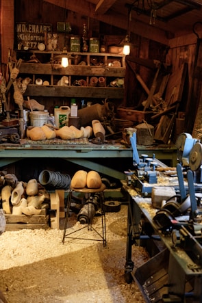 A cluttered workshop filled with woodworking tools and equipment. Wooden clogs are prominently displayed, some resting on a stool in the center. Shelving is packed with various items, including cans, jars, and wooden molds. The floor is covered in sawdust, adding to the rustic atmosphere. Overhead lighting provides a warm glow to the scene.