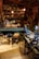 A cluttered workshop filled with woodworking tools and equipment. Wooden clogs are prominently displayed, some resting on a stool in the center. Shelving is packed with various items, including cans, jars, and wooden molds. The floor is covered in sawdust, adding to the rustic atmosphere. Overhead lighting provides a warm glow to the scene.