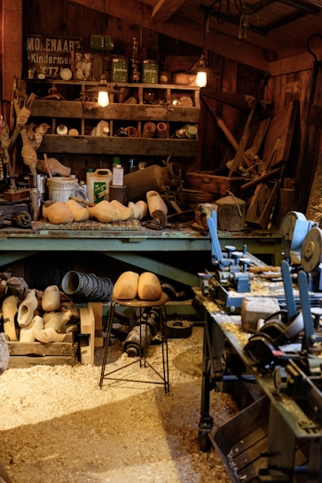 A cluttered workshop filled with woodworking tools and equipment. Wooden clogs are prominently displayed, some resting on a stool in the center. Shelving is packed with various items, including cans, jars, and wooden molds. The floor is covered in sawdust, adding to the rustic atmosphere. Overhead lighting provides a warm glow to the scene.