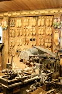 A workshop with an array of wooden clogs hanging on the wall, each pair showing different designs and sizes. In the foreground, a large, worn-out woodcutting machine is covered with wood shavings, indicating active use for crafting the clogs. The environment is rustic, with wooden walls, conveying a traditional craftsmanship setting.