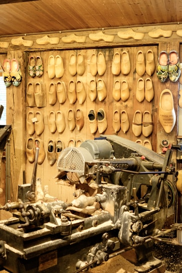 A workshop with an array of wooden clogs hanging on the wall, each pair showing different designs and sizes. In the foreground, a large, worn-out woodcutting machine is covered with wood shavings, indicating active use for crafting the clogs. The environment is rustic, with wooden walls, conveying a traditional craftsmanship setting.