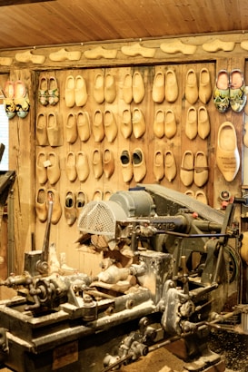 A workshop with an array of wooden clogs hanging on the wall, each pair showing different designs and sizes. In the foreground, a large, worn-out woodcutting machine is covered with wood shavings, indicating active use for crafting the clogs. The environment is rustic, with wooden walls, conveying a traditional craftsmanship setting.