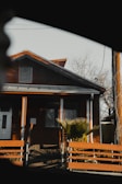 A small wooden house with a gabled roof is partially framed by the interior of a vehicle window. The house has a warm brown exterior with bright orange trim and features a front door and a rectangular window. A palm-like plant is in front of the house, and there is a wooden fence in the foreground. The scene appears to be taken on a clear day, and leafless tree branches are visible in the background.