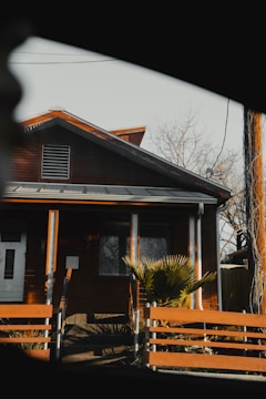 A small wooden house with a gabled roof is partially framed by the interior of a vehicle window. The house has a warm brown exterior with bright orange trim and features a front door and a rectangular window. A palm-like plant is in front of the house, and there is a wooden fence in the foreground. The scene appears to be taken on a clear day, and leafless tree branches are visible in the background.
