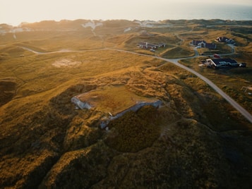 Aerial view of a serene landscape with rolling hills covered in green grass and sandy dunes. Several small houses with slanted roofs are scattered across the scene, connected by narrow dirt paths. The horizon reveals a gentle ocean view under a warm, setting sun that casts a golden hue over the entire area.