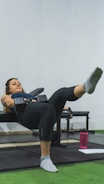 A woman is performing a workout exercise on a bench, holding a dumbbell in each hand while extending one leg outward. She is focused and appears to be in a gym environment with green flooring and gym equipment visible. A pink water bottle and a piece of paper are on the floor nearby.