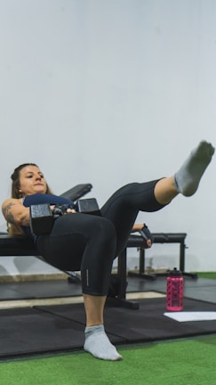 A woman is performing a workout exercise on a bench, holding a dumbbell in each hand while extending one leg outward. She is focused and appears to be in a gym environment with green flooring and gym equipment visible. A pink water bottle and a piece of paper are on the floor nearby.