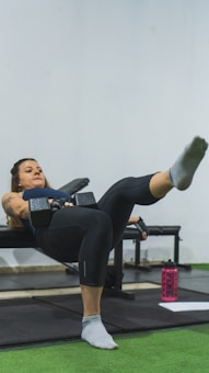 A woman is performing a workout exercise on a bench, holding a dumbbell in each hand while extending one leg outward. She is focused and appears to be in a gym environment with green flooring and gym equipment visible. A pink water bottle and a piece of paper are on the floor nearby.