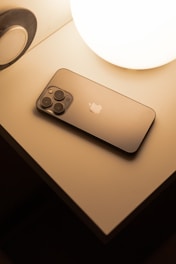 A smartphone with a sleek design lies on a bedside table next to a glowing spherical lamp. The surface of the smartphone is reflective, and the soft lighting creates a warm atmosphere.