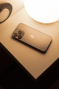 A smartphone with a sleek design lies on a bedside table next to a glowing spherical lamp. The surface of the smartphone is reflective, and the soft lighting creates a warm atmosphere.