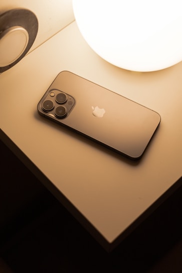 A smartphone with a sleek design lies on a bedside table next to a glowing spherical lamp. The surface of the smartphone is reflective, and the soft lighting creates a warm atmosphere.