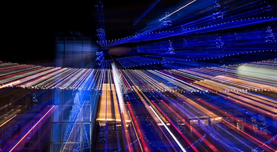 A visually dynamic scene featuring streaks of colorful lights radiating across the image, creating a sense of movement and energy. The predominant colors are blue with accents of red and white, suggesting a festive or celebratory atmosphere. The background includes architectural elements that are partially visible through the light trails.
