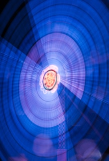 An abstract image featuring concentric circular patterns with a central light source emitting white and orange hues. The dominant color is a vibrant blue, complemented by purple and pink shades that radiate outward in a dynamic, swirling motion.