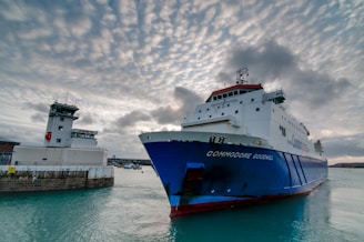 A large cargo ship named 'Commodore Goodwill' is sailing near a harbor. The ship is painted blue and white, with a distinct red area on the top. A nearby harbor control tower is visible to the left, and the sky is filled with dramatic, cloud-laden patterns, adding texture to the scene.