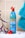 A blue bottle of natural shampoo featuring keratin and ginseng is placed on a tiled surface. The bottle is adorned with a floral design and is topped with a red flower. A turquoise towel is positioned to the right, and a purple leafy branch with red petals lies to the left.
