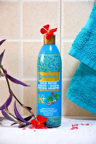 A blue bottle of natural shampoo featuring keratin and ginseng is placed on a tiled surface. The bottle is adorned with a floral design and is topped with a red flower. A turquoise towel is positioned to the right, and a purple leafy branch with red petals lies to the left.