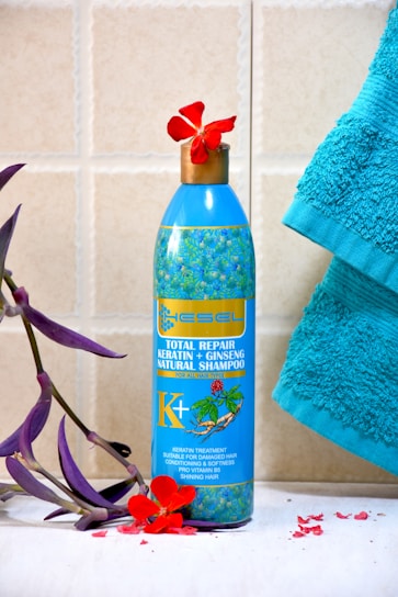 A blue bottle of natural shampoo featuring keratin and ginseng is placed on a tiled surface. The bottle is adorned with a floral design and is topped with a red flower. A turquoise towel is positioned to the right, and a purple leafy branch with red petals lies to the left.