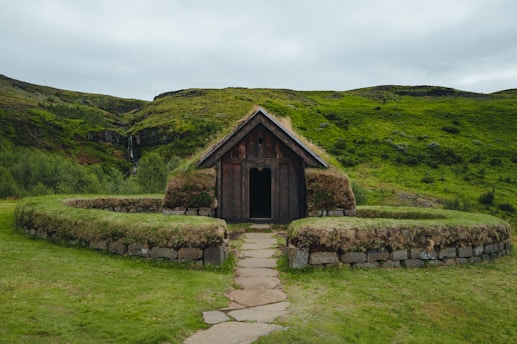 A small, rustic building with a wooden structure and a grass-covered roof sits in the foreground, surrounded by lush green hills. The building is framed by an arched stone pathway and earthwork walls that are also topped with grass. A waterfall in the distance enhances the natural setting, contributing to the serene landscape.
