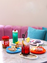 A colorful assortment of foods and beverages is displayed on a table with a marble surface. There are different plates with desserts, including cookie dough and pastries, a jar of Lotus Biscoff spread, and two vibrant drinks, one blue with a cherry on top and one red with a lemon slice. The background features a pink cushioned bench and a light blue cushion.
