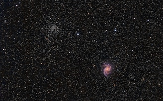 A vast, star-filled cosmos with a bright spiral galaxy prominent against a dark backdrop. Countless stars of varying intensities and colors are scattered throughout, with clusters and formations visible. The galaxy displays pink, purple, and blue hues in its spiral arms.