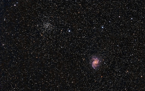 A vast, star-filled cosmos with a bright spiral galaxy prominent against a dark backdrop. Countless stars of varying intensities and colors are scattered throughout, with clusters and formations visible. The galaxy displays pink, purple, and blue hues in its spiral arms.