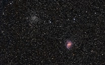 A vast, star-filled cosmos with a bright spiral galaxy prominent against a dark backdrop. Countless stars of varying intensities and colors are scattered throughout, with clusters and formations visible. The galaxy displays pink, purple, and blue hues in its spiral arms.