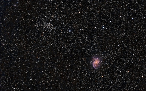 A vast, star-filled cosmos with a bright spiral galaxy prominent against a dark backdrop. Countless stars of varying intensities and colors are scattered throughout, with clusters and formations visible. The galaxy displays pink, purple, and blue hues in its spiral arms.