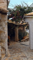 A picturesque stone alleyway with rustic architecture, including stone walls and a tiled roof. Vibrant bougainvillea blooms add color to the scene, while terracotta pots with greenery enhance the quaint, Mediterranean atmosphere.