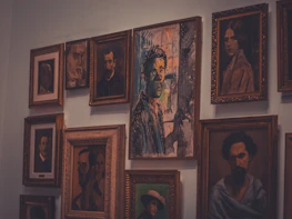 A collection of framed portraits hangs on a wall, featuring a mix of styles from realistic to abstract. The central piece has a striking mix of colors and an expressionistic style.