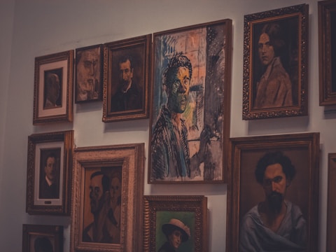 A collection of framed portraits hangs on a wall, featuring a mix of styles from realistic to abstract. The central piece has a striking mix of colors and an expressionistic style.