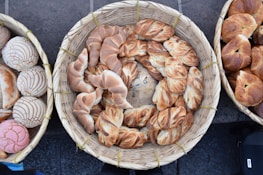A collection of various artisanal bread rolls and pastries is arranged in woven baskets. The breads have distinct textures and appearances, ranging from twisted designs to intricately baked patterns.