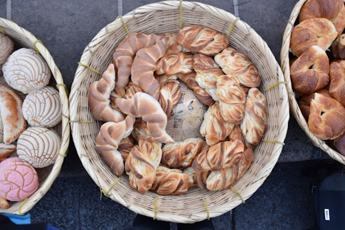 A collection of various artisanal bread rolls and pastries is arranged in woven baskets. The breads have distinct textures and appearances, ranging from twisted designs to intricately baked patterns.