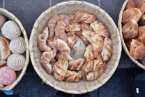 A collection of various artisanal bread rolls and pastries is arranged in woven baskets. The breads have distinct textures and appearances, ranging from twisted designs to intricately baked patterns.