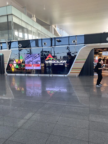 A modern airport terminal features a polished floor and large glass windows. The prominent sign of 'Wroclaw Duty Free' is displayed on a sleek black wall with colorful triangle patterns. A person is walking near the entrance to a shop.