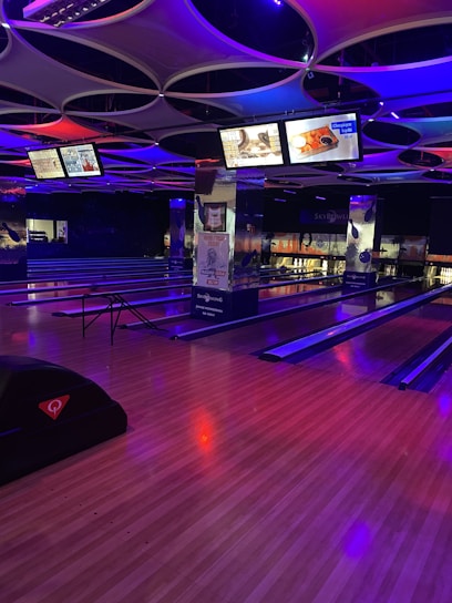 A modern bowling alley with multiple lanes illuminated by colorful neon lights. The ceiling has a futuristic design with circular shapes, and there are digital screens displaying information and advertisements. The wooden floor contrasts with the vivid blue and purple hues from the lighting.