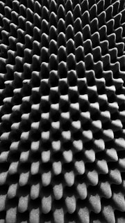 A close-up of a textured surface with a repeating pattern of peaks and valleys, resembling an acoustic foam panel. The surface has a monochrome appearance, showcasing a play of light and shadow across the geometric shapes.