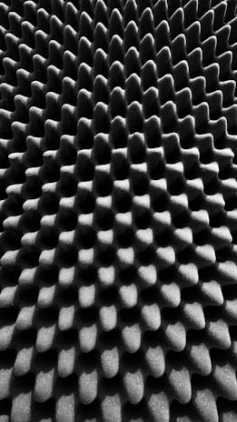 A close-up of a textured surface with a repeating pattern of peaks and valleys, resembling an acoustic foam panel. The surface has a monochrome appearance, showcasing a play of light and shadow across the geometric shapes.