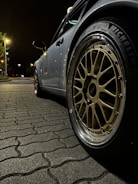 An angled close-up shot of a parked car emphasizes its alloy wheel with a shiny chrome rim. The wheel has distinctive spokes and a visible tire brand marking. The car is situated on a patterned cobblestone pavement under a dimly lit street, with streetlights and faint foliage visible in the background.