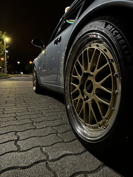 An angled close-up shot of a parked car emphasizes its alloy wheel with a shiny chrome rim. The wheel has distinctive spokes and a visible tire brand marking. The car is situated on a patterned cobblestone pavement under a dimly lit street, with streetlights and faint foliage visible in the background.