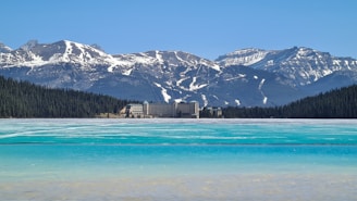 A large turquoise lake is partially frozen, surrounded by dense evergreen forests. In the background, snow-capped mountains rise majestically, highlighting a large, historic hotel building nestled at the shoreline.